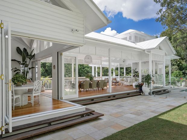 Bright exterior view of a white, pavilion-like residential extension with extensive glazed verandah spaces, louvred and weatherboard-style wall detailing, pitched metal roofs, and a raised timber deck. The covered outdoor dining area opens onto a broad paved terrace, with tropical planting and lawn surrounding the structure.