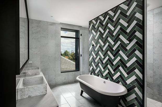 Contemporary bathroom featuring a freestanding black-and-white bathtub, twin square vessel basins, large pale marble-look wall and floor tiles, and a dramatic dark green, white, and grey herringbone feature-tile wall. A black-framed window provides natural light and an exterior view.