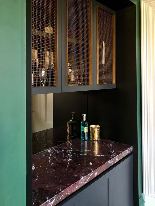A compact, richly detailed built-in bar or drinks cabinet set within a dark-painted alcove. The lower unit has deep burgundy veined stone benchtops and dark grey cabinetry, while the upper storage doors use fine metal mesh panels in dark frames, displaying bottles and glassware. The surrounding walls are painted green, with white decorative door trim visible at the side. The image has a moody, refined, traditional-meets-contemporary feel.