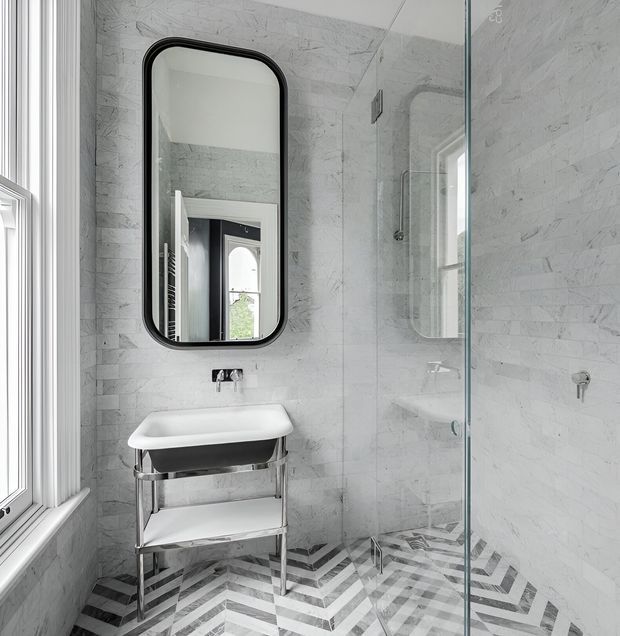 A bright, compact bathroom with pale grey marble-look wall tiles and a bold chevron-patterned tiled floor. The room features a freestanding white basin on a chrome open-frame console, a black-framed rounded-rectangle mirror, a glass shower enclosure, and a white-trimmed window. The design combines crisp contemporary fittings with refined geometric detailing.