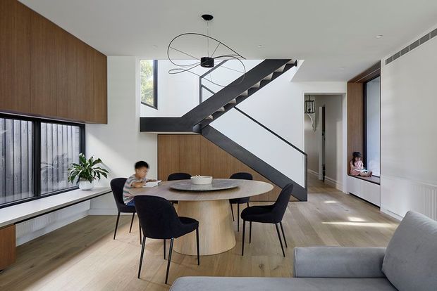 Bright contemporary open-plan dining and living area with a round timber dining table and upholstered chairs, pale timber flooring, built-in timber cabinetry, and a sculptural dark steel staircase with open risers and slim balustrade. Large windows provide abundant natural light, while a grey sofa occupies the foreground and a hallway leads toward another room.