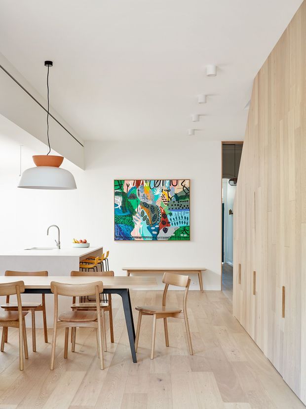 Bright open-plan dining and kitchen interior with pale timber flooring, a white island bench, light timber dining furniture, and a full-height pale timber wall of built-in cabinetry or panelling. A colourful abstract artwork provides a strong focal point, with simple pendant and ceiling lighting creating a clean contemporary atmosphere.