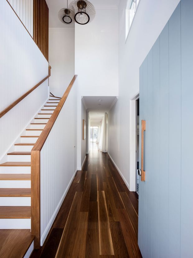 Bright, narrow residential hallway with a timber staircase on the left, white vertical wall panelling, warm dark timber flooring, and pale blue built-in cabinetry or a door with timber handles on the right. Three sculptural black pendant lights hang above the stairwell, while a high window brings in natural light.