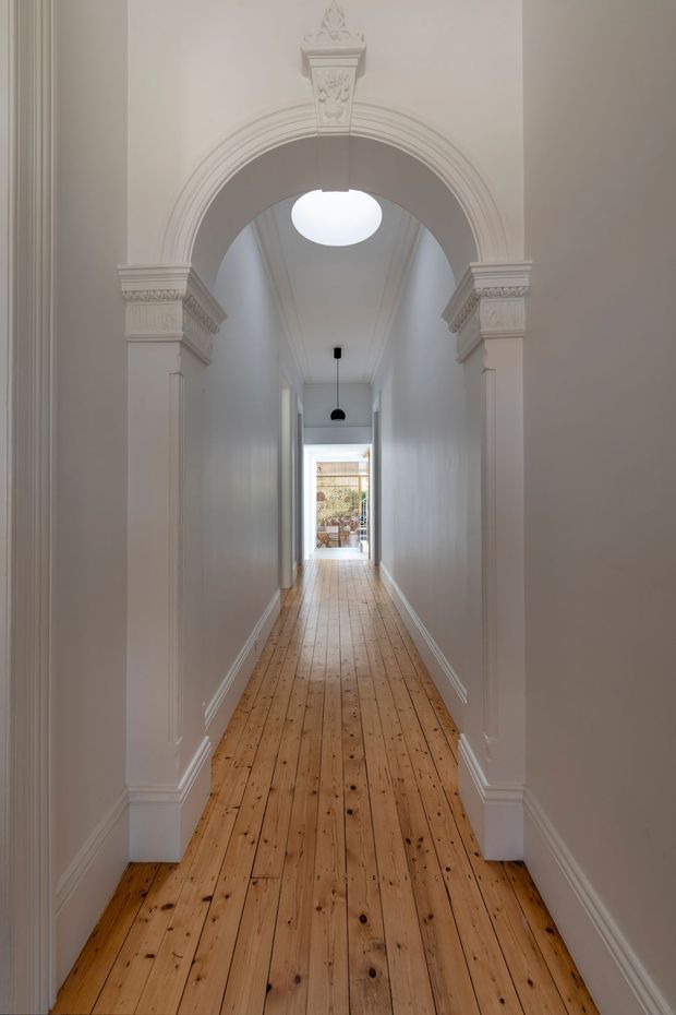 A long, narrow residential hallway with original timber floorboards, white painted walls, ornate decorative architraves, fluted pilasters, and a detailed classical archway. A circular skylight and black pendant light illuminate the corridor, which leads to a glazed doorway and outdoor area at the far end.