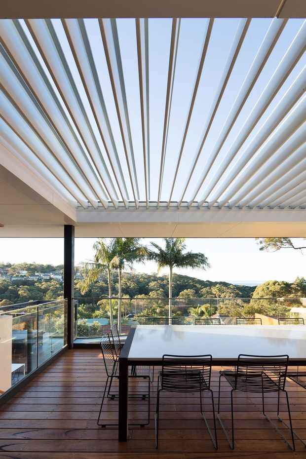 A modern outdoor dining terrace with a long rectangular dining table and black wire-frame chairs on a timber deck. The covered terrace features a dramatic white louvred pergola roof, dark structural posts, glass balustrades, and expansive views over a lush, hilly landscape with palm trees.