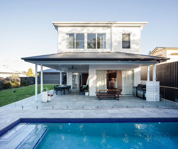 Rear exterior view of a two-storey white weatherboard home with a broad covered outdoor entertaining area opening onto a paved terrace and swimming pool. The facade features extensive glazing, a dark low-pitched metal roof, vertical privacy fencing, lawn, and an outdoor barbecue area.