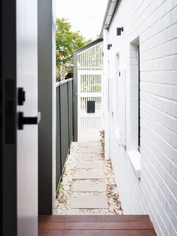 Narrow side passage beside a white-painted brick building, viewed from a dark-framed doorway. The passage has a timber threshold/deck, pale stepping slabs set in white gravel, a dark vertical metal fence, white slatted screening and a small gate at the far end, with simple black wall lights and white rainwater gutters.