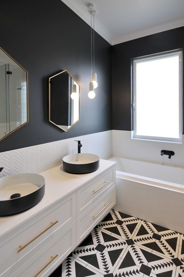 A stylish bathroom featuring a long white vanity with two black vessel basins, black tapware, gold hardware, a white textured herringbone wall tile splashback, and bold black-and-white geometric floor tiles. The room has charcoal walls, white ceiling moulding, a freestanding-style bathtub beneath a large window, and contemporary pendant lighting with geometric mirrors.