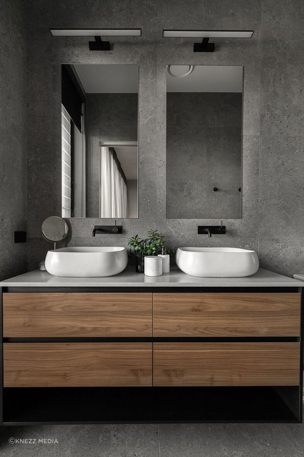 A contemporary double-vanity bathroom with two rectangular mirrors, wall-mounted black faucets, white vessel basins, and a floating timber vanity with dark framing. The walls and floor have a dark textured concrete finish, complemented by minimalist linear mirror lighting and small potted greenery.