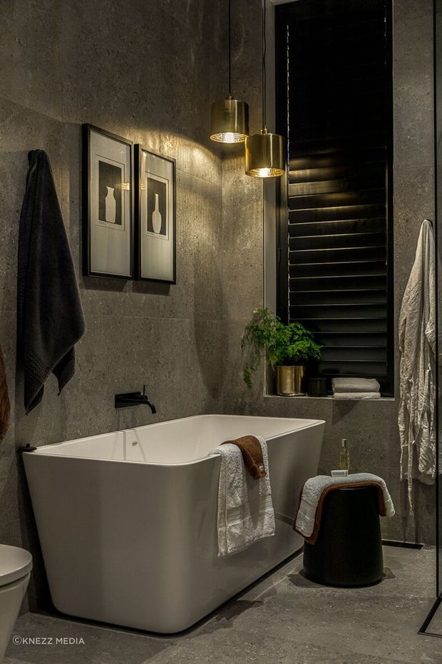 A moody, refined contemporary bathroom featuring a freestanding white bathtub, dark grey concrete-look wall and floor finishes, black window framing and shutters, brushed brass pendant lights, black tapware, framed artwork, towels, and a small stool with a potted plant.