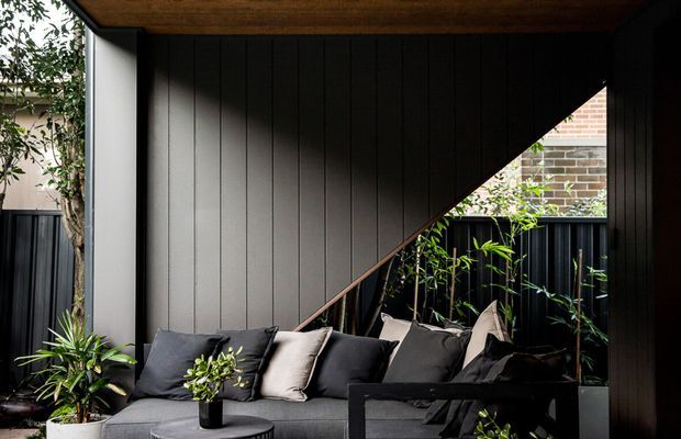 A partially enclosed outdoor lounge with a dark grey sectional sofa, cushions, coffee table, and potted plants. The space is framed by dramatic black vertical wall cladding and a sharply angled timber-lined overhead structure, with glimpses of surrounding fencing and brickwork.
