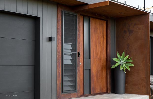 Close-up exterior view of a contemporary residential entry, featuring a large timber pivot door with narrow vertical glazing and a louvred sidelight. The entry is framed by dark grey vertical cladding, a projecting timber soffit, and a rust-toned metal privacy screen, with a tall black planter beside the door. A flush dark garage door is visible at left.