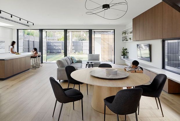 Open-plan contemporary living and dining area with a light timber floor, sculptural round dining table and upholstered black dining chairs in the foreground. A pale sectional sofa faces a wall-mounted television and built-in timber cabinetry, while a kitchen island and stools sit to the left. Full-height glazed doors open toward a private courtyard, and a large linear track light and sculptural ceiling pendant provide modern lighting.