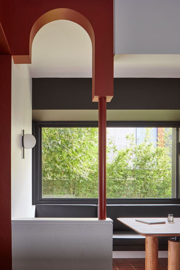 A striking contemporary dining nook with a built-in black banquette beneath large dark-framed windows, overlooking dense greenery. The room features bold deep red architectural columns and trim, including a decorative arch, white plaster walls, a sculptural wall sconce, and a light timber dining table with rounded legs. Terracotta-toned floor tiles and strong geometric contrasts create a refined, graphic interior.