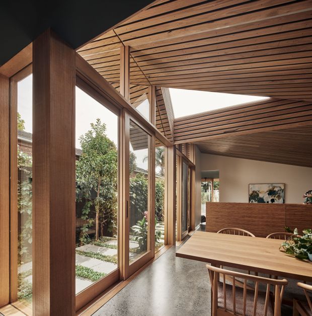 A warm, contemporary dining area within a glazed indoor-outdoor corridor. Full-height timber-framed glazing overlooks a planted courtyard, while a slatted timber ceiling continues across the room and features a large skylight opening. A timber dining table and chairs sit beside a dark polished concrete floor, with a timber room divider and kitchen or living area beyond.