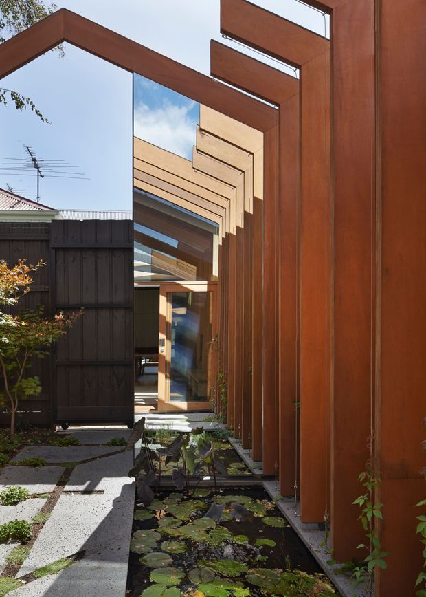 A narrow landscaped courtyard passage beside a contemporary timber-clad building. Deep reddish-brown vertical timber panels and projecting timber frames create a strong rhythmic facade, with extensive glazing and a recessed entry at the end of the passage. A shallow pond with lily pads runs alongside a concrete stepping-stone path, while a dark timber privacy fence and neighbouring roof are visible beyond. Bright daylight, strong shadows, and reflections emphasize the layered architectural composition.