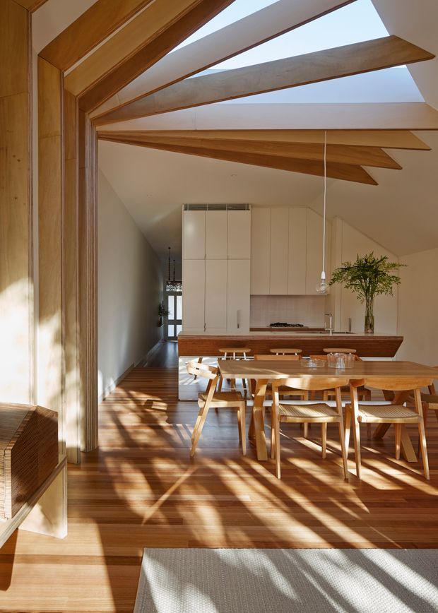 A sunlit open-plan dining and kitchen interior with extensive warm timber detailing. A large timber dining table and matching chairs sit on timber flooring, while exposed timber roof beams and a sloped ceiling create a strong structural feature overhead. The rear kitchen has streamlined off-white cabinetry, a timber island or counter front, and a pale tiled backsplash. Strong daylight casts geometric shadows across the floor and furniture.