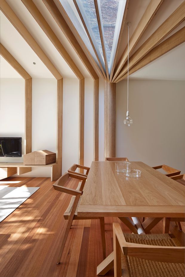 A warm, light-filled dining and living area with a large solid timber dining table and matching timber-framed chairs. Exposed timber ceiling beams and vertical wall ribs radiate toward a central roof skylight, creating a strong structural feature. Timber flooring, white plaster walls, a simple pendant light, and built-in low cabinetry with a television complete the refined interior.