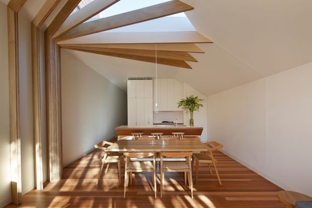 A warm, minimalist open-plan dining and kitchen interior with a timber dining table and chairs in the foreground, opening onto a compact white kitchen. Rich timber flooring, cabinetry, exposed timber ceiling beams, and large roof glazing create strong linear shadows and a bright, sunlit atmosphere.