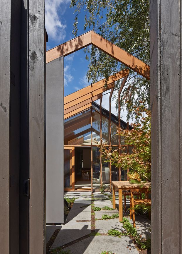 A narrow architectural view through dark timber-clad walls into a sunlit courtyard and glazed building entrance. The composition highlights exposed warm timber beams forming a pitched roof structure, extensive floor-to-ceiling glazing, concrete stepping slabs, planted courtyard beds, and a timber outdoor table. Mature trees and blue sky are visible beyond the open structure.