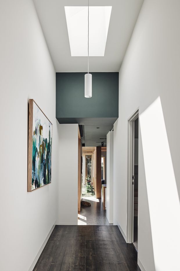 A bright, narrow residential hallway with white walls, dark timber flooring, and a high ceiling featuring a large rectangular skylight and a simple cylindrical pendant light. A dark charcoal ceiling bulkhead creates a strong visual transition toward the far end, where timber screens, glazing, and a glimpse of an adjoining living area are visible. An abstract painting hangs on the left wall, while doorways line the right side. Strong sunlight creates geometric shadows on the walls.