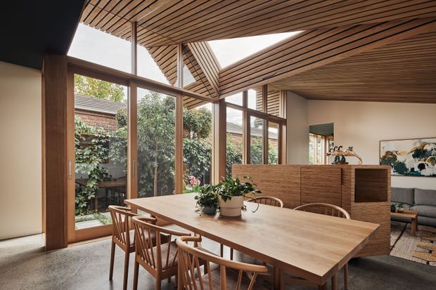 A warm, light-filled dining area with a large timber dining table and wooden chairs, surrounded by floor-to-ceiling glazing opening toward a planted courtyard. The room features an exposed timber slatted ceiling with angled structural detailing, timber-framed windows and doors, polished concrete flooring, and a timber built-in divider separating the dining space from a partially visible living room.