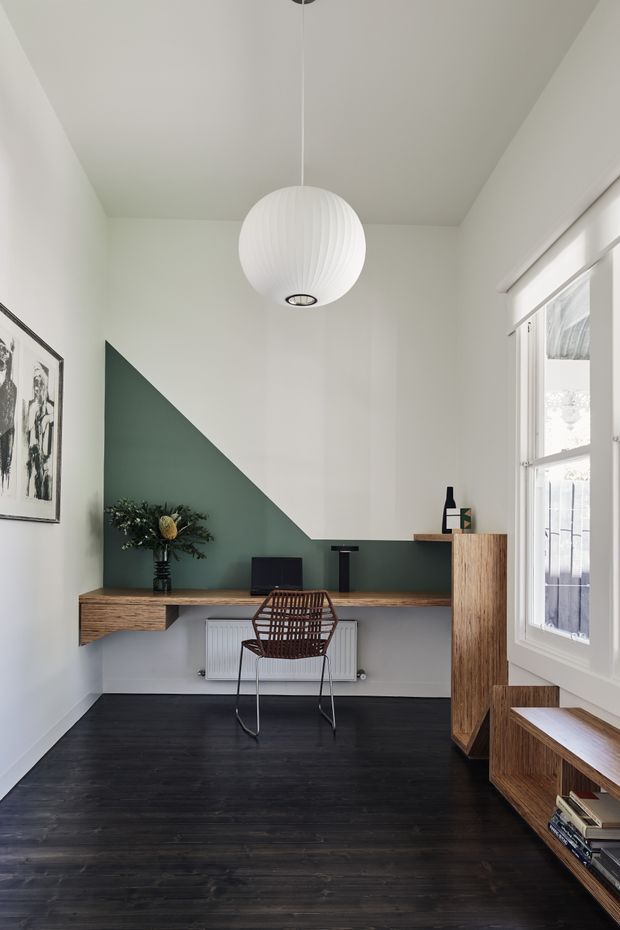 A compact, bright home office with white walls and ceiling, a dark green geometric feature wall, and a long floating timber desk. The room includes a woven wire chair, black computer monitor, sculptural white pendant light, timber storage cabinetry and low shelving, framed artwork, and dark timber flooring. Tall white-framed windows provide natural light.