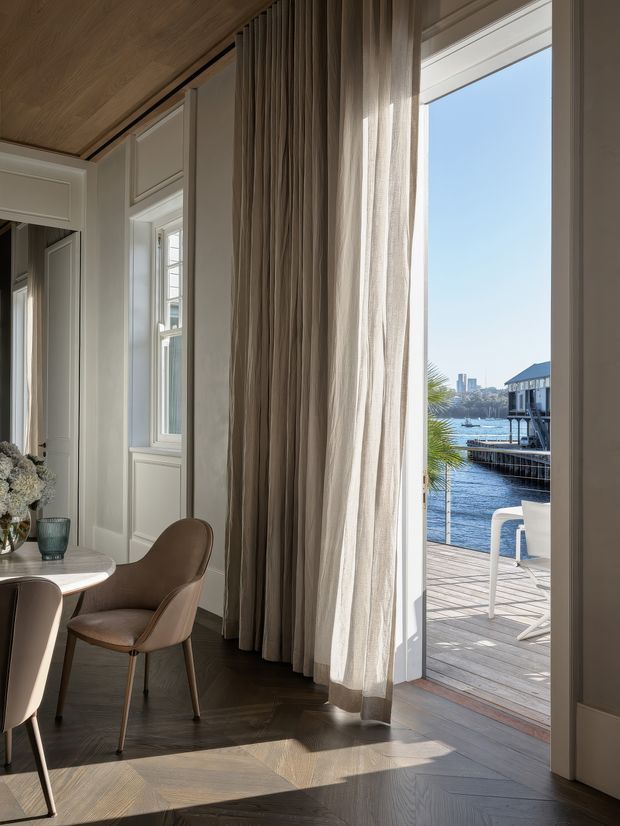 A refined dining area opening onto a waterfront timber deck. The room features pale panelled walls and trim, full-height sheer and pleated taupe curtains, a dark herringbone timber floor, a small round dining table, and upholstered brown dining chairs. Sunlight enters through tall windows and an open glazed door, with water, a palm, and neighbouring waterfront structures visible outside.