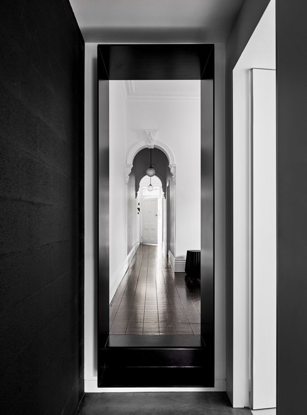 A monochromatic architectural photograph looking through a tall, black-framed interior opening or mirror toward a long, narrow hallway. The hallway has white walls, ornate arched door surrounds, pendant lights, a white panelled door at the far end, and dark timber flooring. The foreground is highly graphic and dark, with black wall and framing contrasted against bright white door jambs and corridor surfaces.