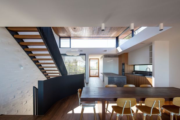 Open-plan contemporary home interior showing a dining area in the foreground, kitchen beyond, and a sculptural open-riser staircase with black metal balustrade. The space has a warm timber ceiling and flooring, white-painted brick walls, dark stone kitchen surfaces, extensive glazing, and a double-height void with clerestory-style light.
