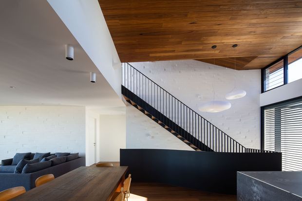 Open-plan contemporary interior showing a dining area in the foreground, a living room to the left, and a black metal stair with a solid privacy balustrade leading to an upper level. The space features white-painted brick walls, a warm timber-lined ceiling, dark timber flooring, large windows with horizontal blinds, and sculptural pendant lighting.