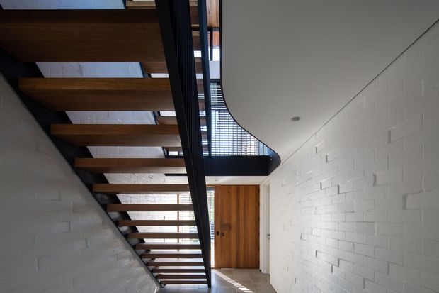 A modern interior stairway with open timber treads and dark metal stringers, rising alongside white-painted brick walls. A curved black balustrade and upper landing overlook the stair, while a timber door and narrow glazing bring warm natural light into the otherwise minimal circulation space.
