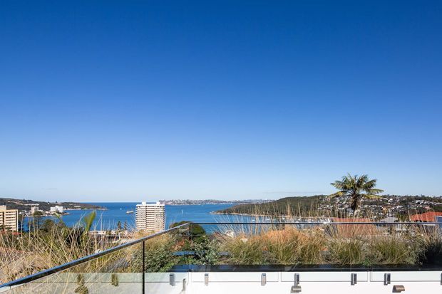 Elevated exterior terrace or balcony overlooking a coastal bay, with a transparent glass balustrade, landscaped ornamental grasses, and a broad urban waterfront view beneath a clear blue sky.