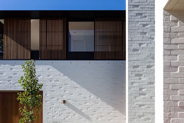 Exterior architectural photograph of a contemporary multi-level facade. The building features white-painted brick walls, dark timber vertical screening across upper-level openings, a deep dark roof or fascia, and a timber entry door. Strong sunlight creates geometric shadows across the masonry, with a small planted tree in the foreground.