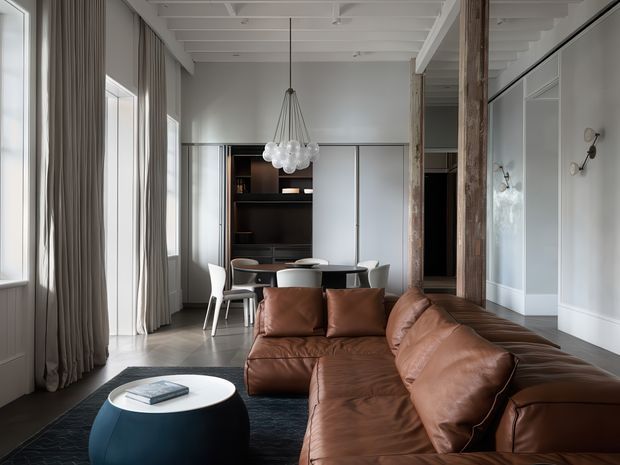 A refined open-plan living and dining area with a large caramel-brown leather sectional sofa in the foreground, a round dark coffee table, and a compact dining setting behind it. Tall windows with full-length curtains line the left wall, while exposed timber columns, pale wall panelling, and a white painted coffered or beam ceiling define the architecture. A sculptural glass pendant hangs above the dining table, and built-in dark joinery provides storage at the rear.