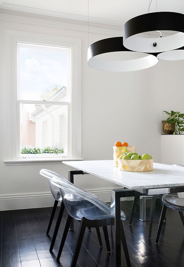 Bright, refined dining area with a white marble-look rectangular dining table, transparent acrylic chairs with dark legs, dark timber flooring, pale walls, traditional decorative wall mouldings, and a large black-and-white circular pendant light. A window brings in natural light and a small potted plant sits on a wall shelf.