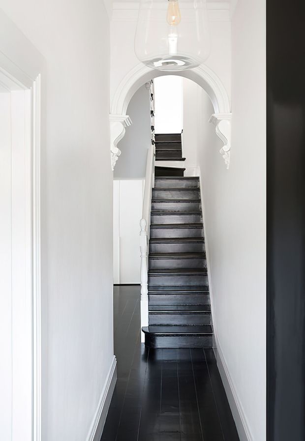 A narrow, bright residential hallway focused on a traditional staircase. The staircase has dark painted timber treads and risers, a white turned newel post and balustrade, and rises through an ornate white arched opening with decorative corbels. Glossy dark flooring contrasts with the white walls and trim, while a clear glass pendant light hangs overhead.