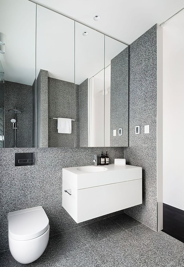 A compact contemporary bathroom with a wall-hung white vanity and integrated basin, wall-mounted toilet, large mirrored cabinet, and shower visible in the mirror. Grey-and-white speckled tile covers the walls and floor, with recessed ceiling lights and minimal chrome fixtures.