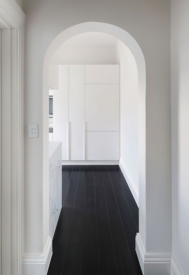 A narrow, bright transitional hallway viewed through a prominent white arched opening. The corridor has dark timber-look plank flooring, white painted walls, decorative skirting and door trim, and minimalist full-height white built-in cabinetry at the far end. A partial kitchen or utility area is visible to the left beyond the cabinetry.