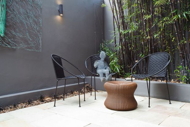 A compact landscaped courtyard or outdoor seating area with pale stone paving, dark rendered walls, black wire-frame lounge chairs, and a woven brown sculptural side table. Tall bamboo planting creates a private green screen, with a small wall light and decorative artwork adding visual interest.