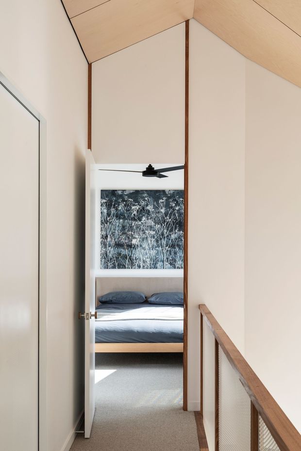 A narrow upper-level hallway or landing looks through an open doorway into a calm, minimalist bedroom. The corridor has a sloped light timber ceiling, white walls, dark timber trims, carpeted flooring, and a timber-framed mesh balustrade. The bedroom beyond contains a low timber bed with blue-grey bedding, a black ceiling fan, and a large monochrome botanical artwork.