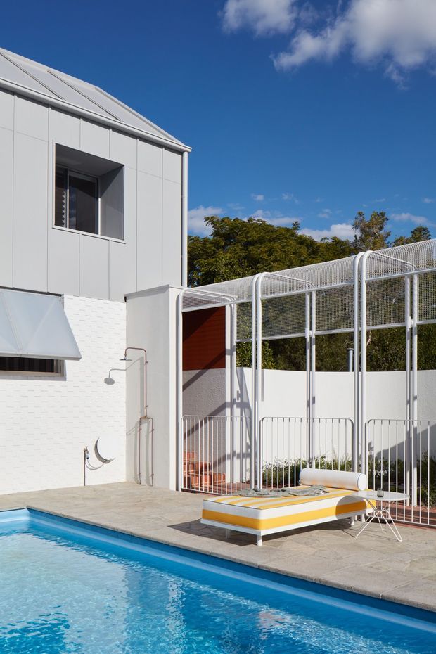 Bright exterior view of a contemporary white-clad building beside a swimming pool. The scene includes a paved pool deck, a white metal and mesh-covered outdoor structure, a sculptural yellow-and-white sun lounger, and a small outdoor shower. Strong blue sky and mature trees form the background.