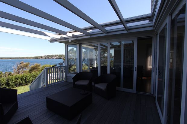 A covered waterfront deck or terrace with dark timber decking, a pergola-style overhead structure, glass sliding doors and extensive glazing. Dark outdoor lounge chairs and a low coffee table overlook a calm body of water and the opposite shoreline.