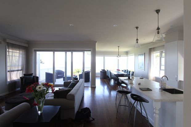 Open-plan coastal-style home interior combining a living room, dining area, and kitchen. The space has dark timber flooring, white cabinetry and walls, a long white kitchen island with dark benchtop, pendant lighting, large glazed doors and windows, and direct views toward a bright outdoor setting. A sectional sofa, dining table, bar stools, and kitchen fixtures are visible.