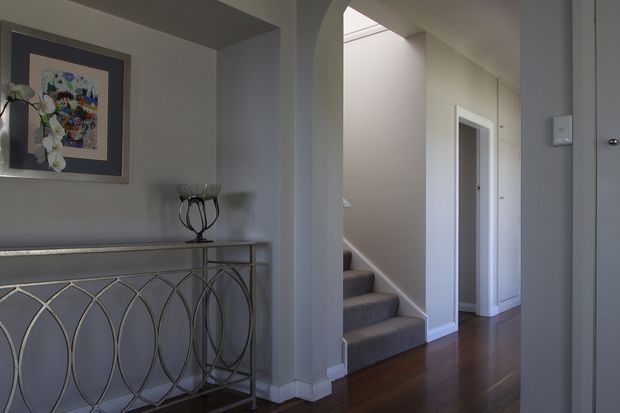 A refined residential hallway and entry area with grey walls, dark timber flooring, and a carpeted staircase rising through an arched opening. A decorative console with a geometric metal base sits beneath framed artwork, while white-trimmed doorways and a skylit stairwell add architectural interest.