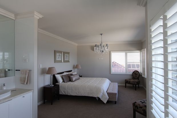 A bright, refined bedroom viewed from an adjoining bathroom or dressing area. The room features a neatly made upholstered bed with layered neutral bedding, matching bedside lamps, framed artwork, a buttoned bench at the foot of the bed, a chandelier, carpeted flooring, and plantation shutters over the windows. A vanity and glass shower enclosure are visible at the left edge, indicating a direct connection to the ensuite.