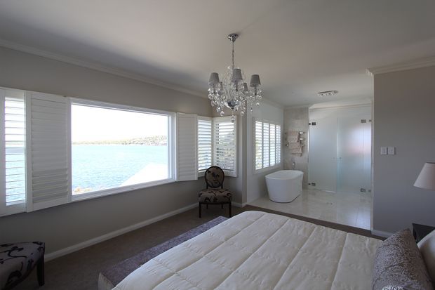 Bright, elegant bedroom overlooking a body of water, with a large window fitted with white plantation shutters, a tufted bed, crystal chandelier, occasional chair, and an open ensuite bathroom featuring a freestanding white bathtub and glass shower enclosure. The room has neutral walls, pale tiled bathroom flooring, and refined traditional detailing.