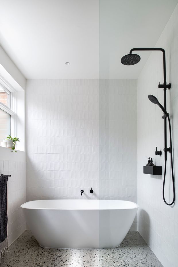 Bright, compact contemporary bathroom featuring a freestanding white bathtub, black wall-mounted rain shower and handheld shower, white textured wall tiles, terrazzo-style flooring, and a large window with a small potted plant. The space has a clean, minimal, high-contrast monochrome finish.