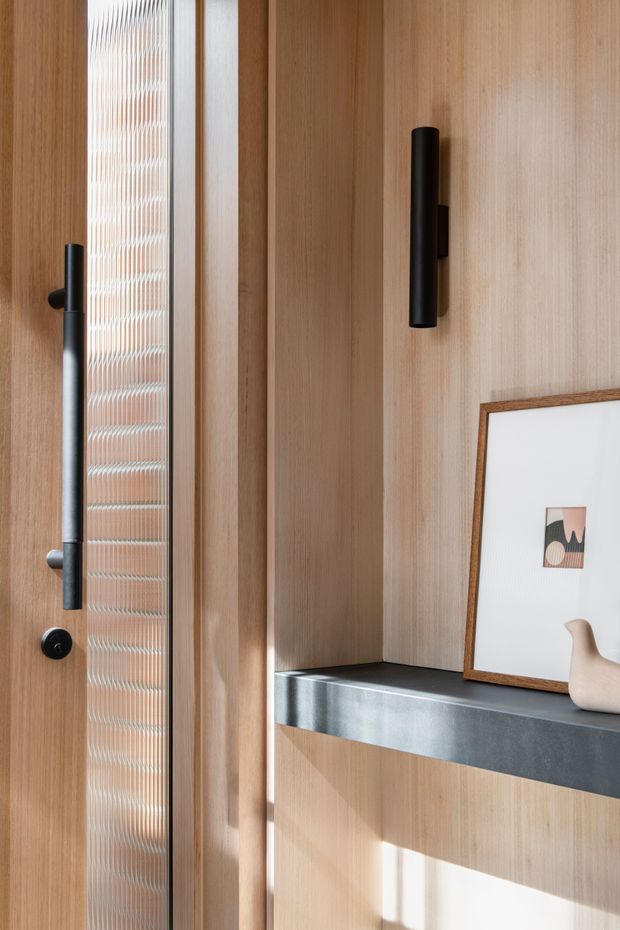 Close-up architectural interior detail showing a timber-clad entryway with a tall reeded-glass door, black cylindrical door hardware, integrated timber wall panels, and a dark stone or engineered-stone shelf. Warm natural light highlights the refined minimalist joinery. A framed artwork and small sculptural object sit on the shelf.