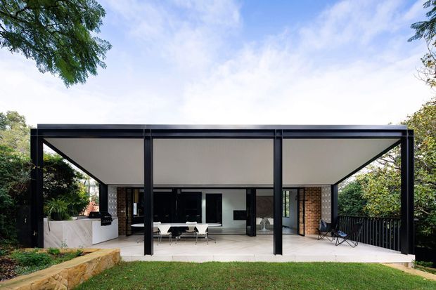Exterior view of a contemporary single-storey pavilion-style home with a broad flat roof, black metal framing, pale soffit, extensive open glazing, brick walls and a covered outdoor entertaining area. The covered terrace includes dining and lounge furniture, while landscaped greenery surrounds the building.